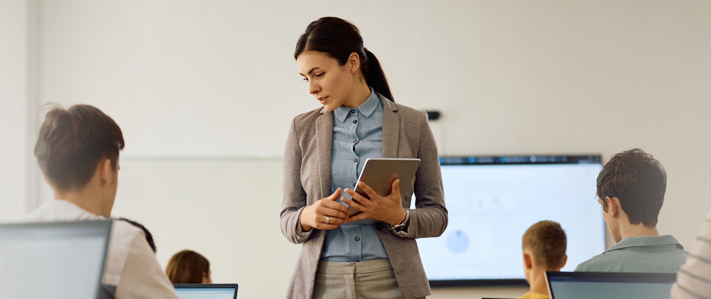 Woman holds table and observes students on laptops.