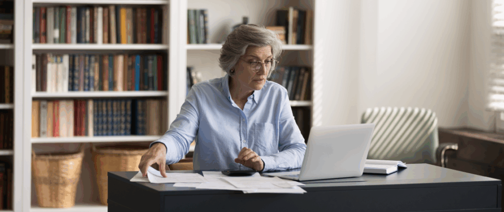 Senior female professor with gray hair working on laptop reviewing papers in home office with bookshelves