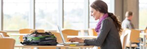 Female college student studying on laptop in bright university library with backpack and textbooks