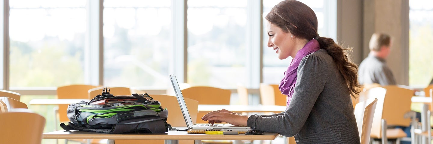 Female college student studying on laptop in bright university library with backpack and textbooks