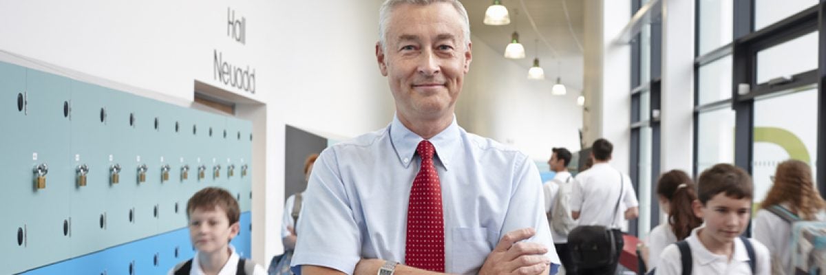 Smiling K-12 administrator with arms crossed stands by lockers in school hallway as young students head to class