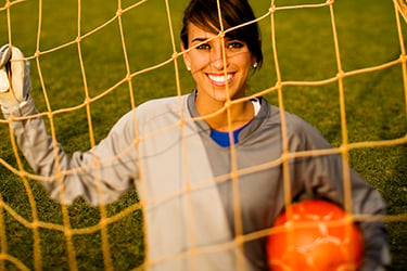 Athlete holding a soccer ball on a field behind a goal net.